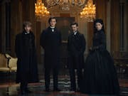 Three young white men in black suits and one white woman in black dress and hat stand in a grand house with crystal chandeliers
