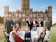 A large group of people in 1930s period clothing sitting in front of a grand house and estate