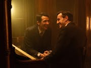 Two young white men in suits sitting at a piano laughing and leaning into each other