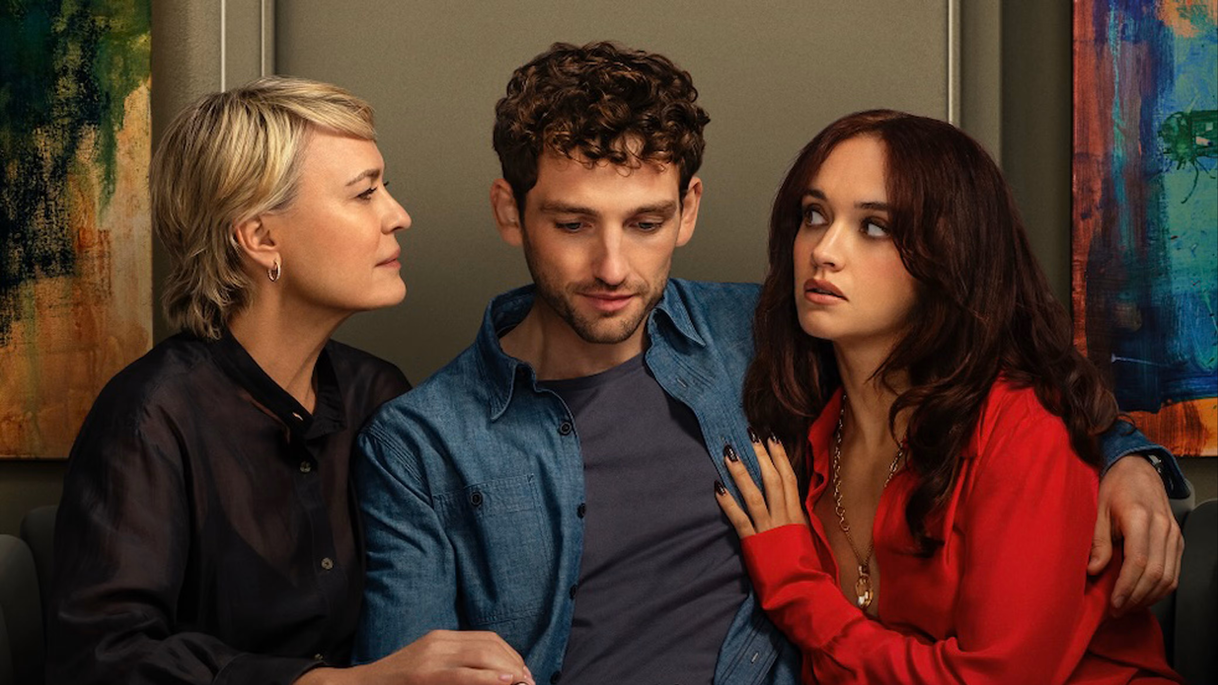 A middle aged woman sits possessively beside a young man who has his arm around a young woman as the women stare at each in a standoff