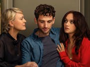 A middle aged woman sits possessively beside a young man who has his arm around a young woman as the women stare at each in a standoff