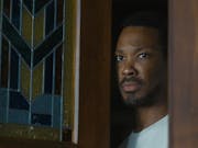 A Black man standing in his dark wood and stain glass window doorway looking cautious