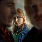 A red haired woman is looking at the camera. In from of her, two out of focus men talking to each other