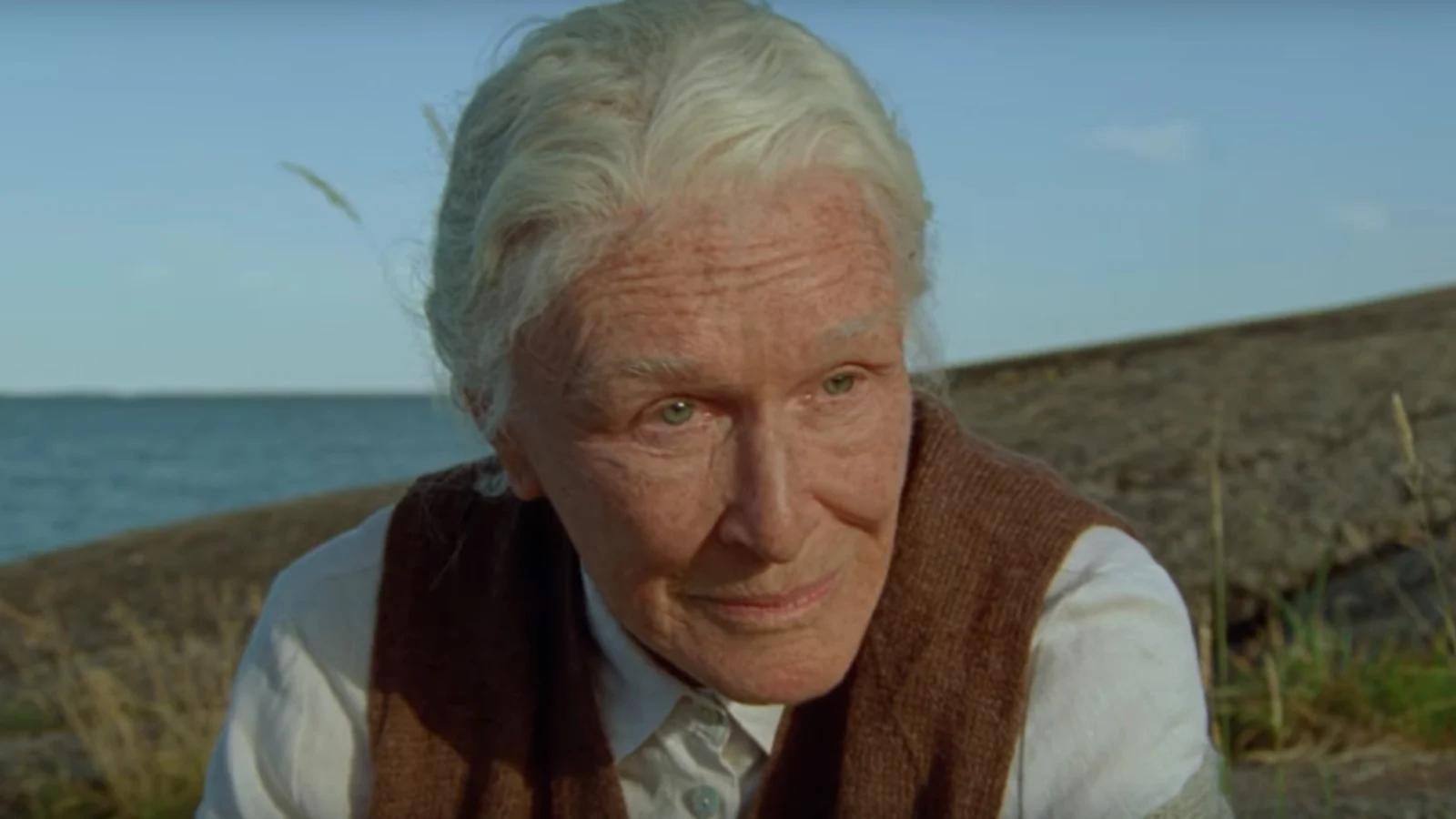 white woman with white hair looking toward the camera, behind her the sea.
