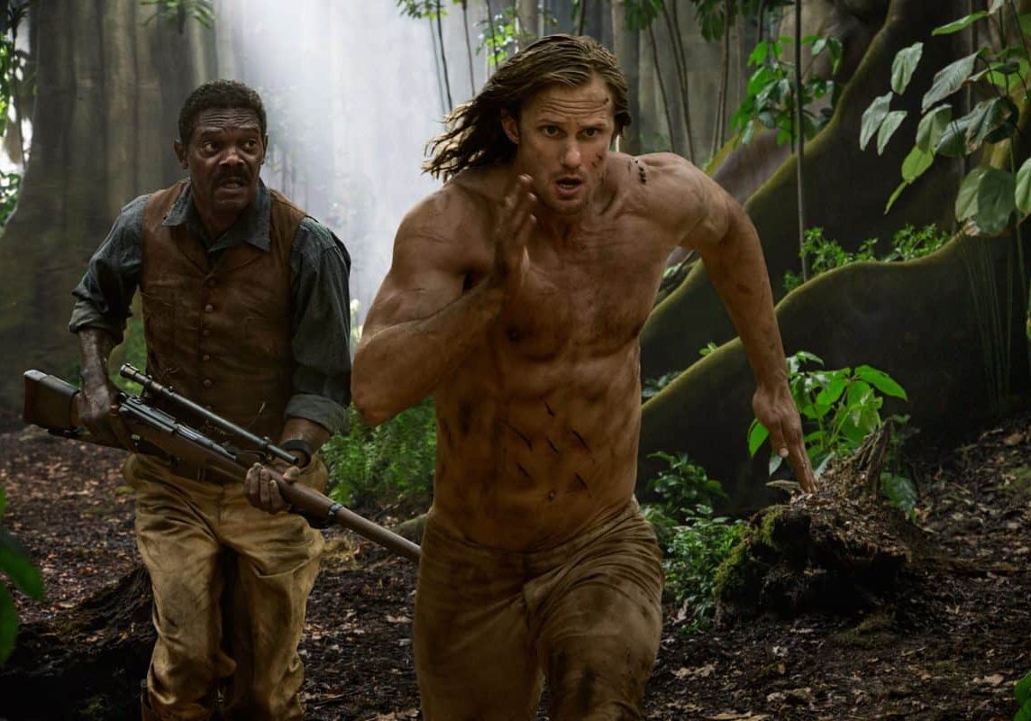 A white man with long hair and a black man with a moustache run in the jungle. the black man is holding a rifle