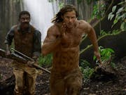 A white man with long hair and a black man with a moustache run in the jungle. the black man is holding a rifle