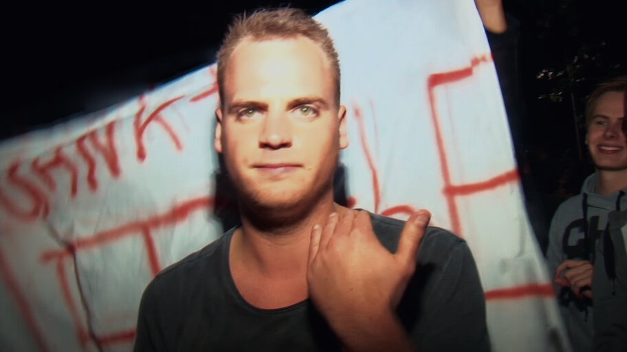 A white man looking at the camera in front of a banner with red letters