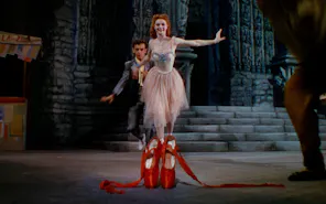 A red-haired woman wearing a pink fairy-like dress, running towards a pair of red ballet slippers in the foreground. Behind her, a crouched man, eyeing the shoes, chasing after her