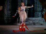 A red-haired woman wearing a pink fairy-like dress, running towards a pair of red ballet slippers in the foreground. Behind her, a crouched man, eyeing the shoes, chasing after her