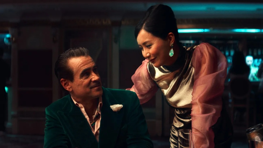 A middle aged white man in a green velvet suit sits at a poker table, smiling at a Chinese woman in an elegant pink dress hovering beside him