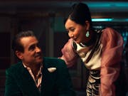 A middle aged white man in a green velvet suit sits at a poker table, smiling at a Chinese woman in an elegant pink dress hovering beside him