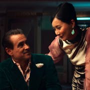 A middle aged white man in a green velvet suit sits at a poker table, smiling at a Chinese woman in an elegant pink dress hovering beside him
