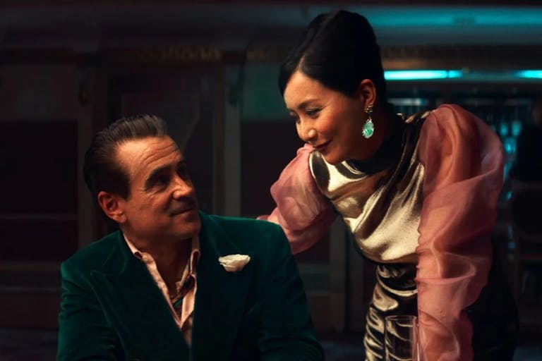 A middle aged white man in a green velvet suit sits at a poker table, smiling at a Chinese woman in an elegant pink dress hovering beside him
