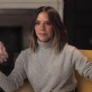 A white woman with a bob and beige turtleneck jumper sits elegantly in a chair beside a vase of pink flowers giving a talking head interview
