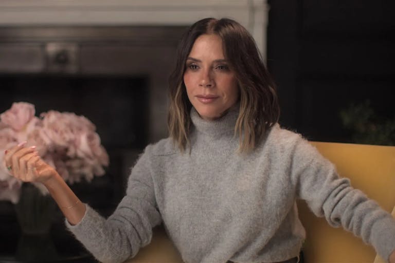 A white woman with a bob and beige turtleneck jumper sits elegantly in a chair beside a vase of pink flowers giving a talking head interview