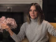 A white woman with a bob and beige turtleneck jumper sits elegantly in a chair beside a vase of pink flowers giving a talking head interview