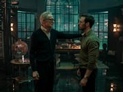 A middle aged white man wearing glasses and a cardigan stands with his hand on the should of a young white man