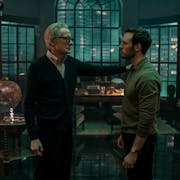 A middle aged white man wearing glasses and a cardigan stands with his hand on the should of a young white man