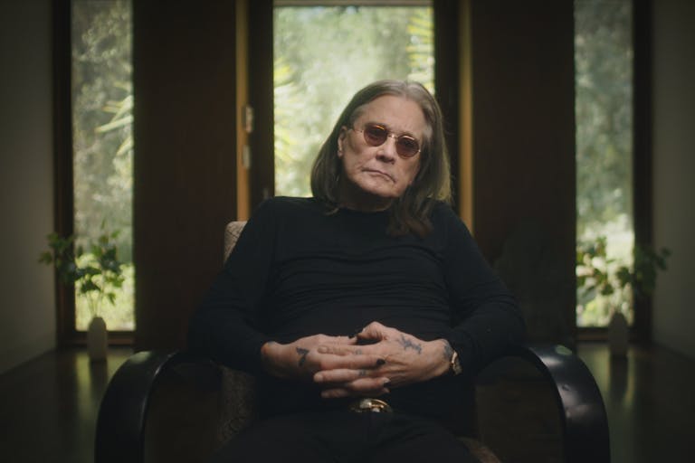 An old white man with long grey and dark hair and black clothing, rings and tattoos on his hand, wearing small round dark glasses, sits in a chair giving a talking head interview