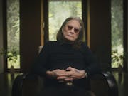 An old white man with long grey and dark hair and black clothing, rings and tattoos on his hand, wearing small round dark glasses, sits in a chair giving a talking head interview