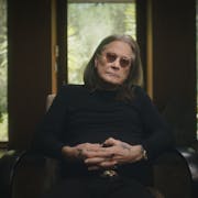 An old white man with long grey and dark hair and black clothing, rings and tattoos on his hand, wearing small round dark glasses, sits in a chair giving a talking head interview