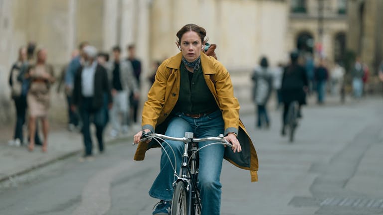 A white woman on a yellow raincoat and blue jeans cycles down a busy street