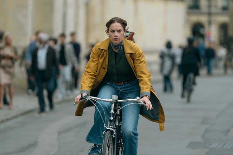 A white woman on a yellow raincoat and blue jeans cycles down a busy street