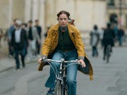 A white woman on a yellow raincoat and blue jeans cycles down a busy street