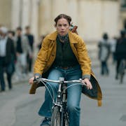 A white woman on a yellow raincoat and blue jeans cycles down a busy street