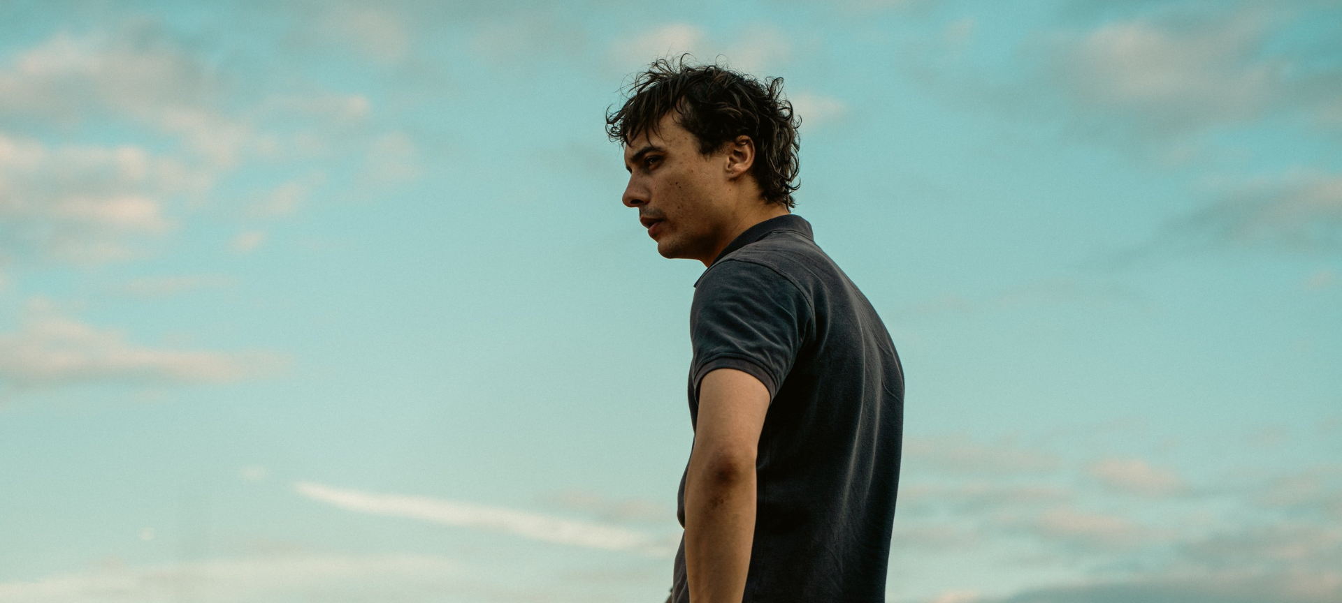 A young slim white man with messy dark hair looks off into the distance with a bright blue sky behind him