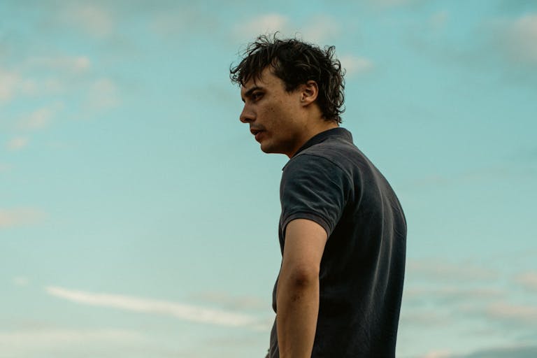 A young slim white man with messy dark hair looks off into the distance with a bright blue sky behind him