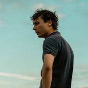 A young slim white man with messy dark hair looks off into the distance with a bright blue sky behind him