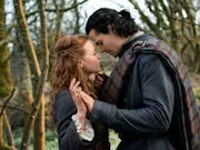 A man and a woman embracing in the woods. The man is wearing dark tartan