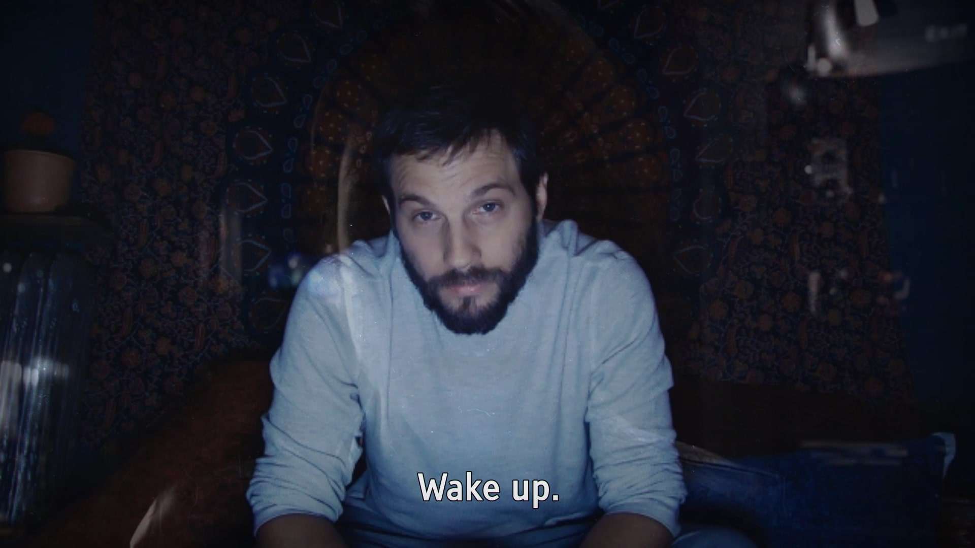 A bearded white man wearing a white t-shirt sits in a dark room looking at the camera. Subtitles read 'Wake up'.