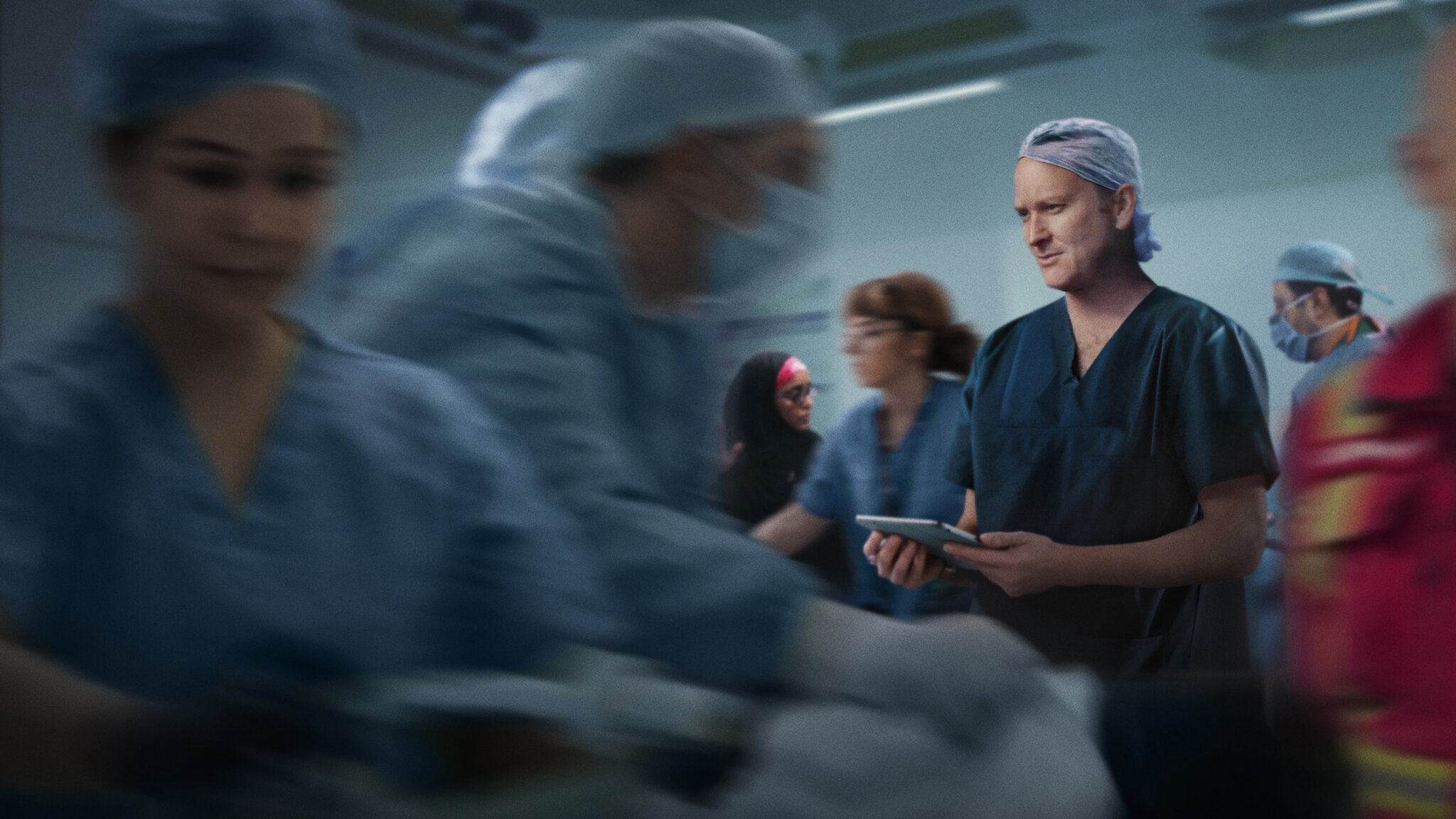 A group of people wearing scrbus prepare for surgery. Only one of them is in focus and the rest appear to be moving very fast