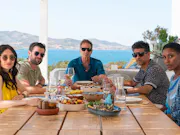 A small group of people sitting around a table in a warm, bright holiday destination with clear blue seas behind them