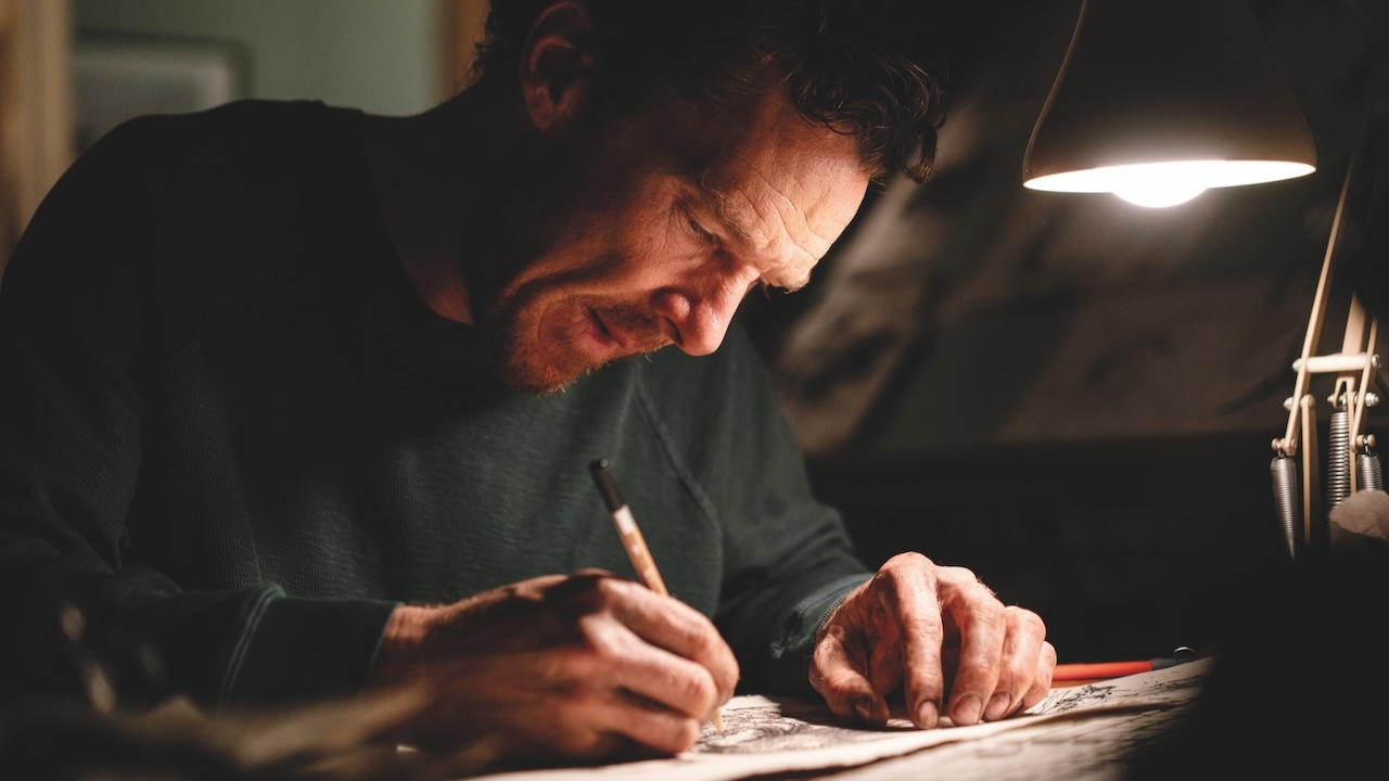 A white man sits at a desk with a low lamp on scribbling with a pencil
