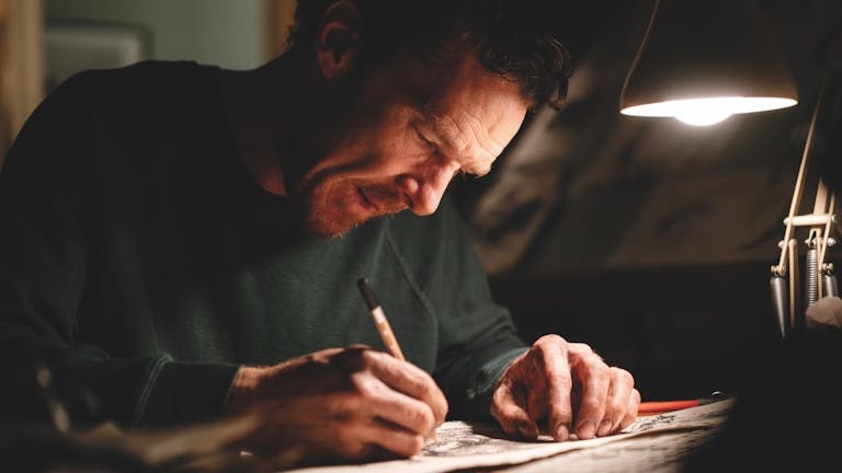 A white man sits at a desk with a low lamp on scribbling with a pencil 
