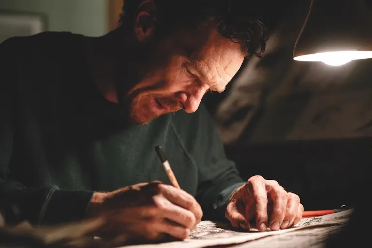 A white man sits at a desk with a low lamp on scribbling with a pencil