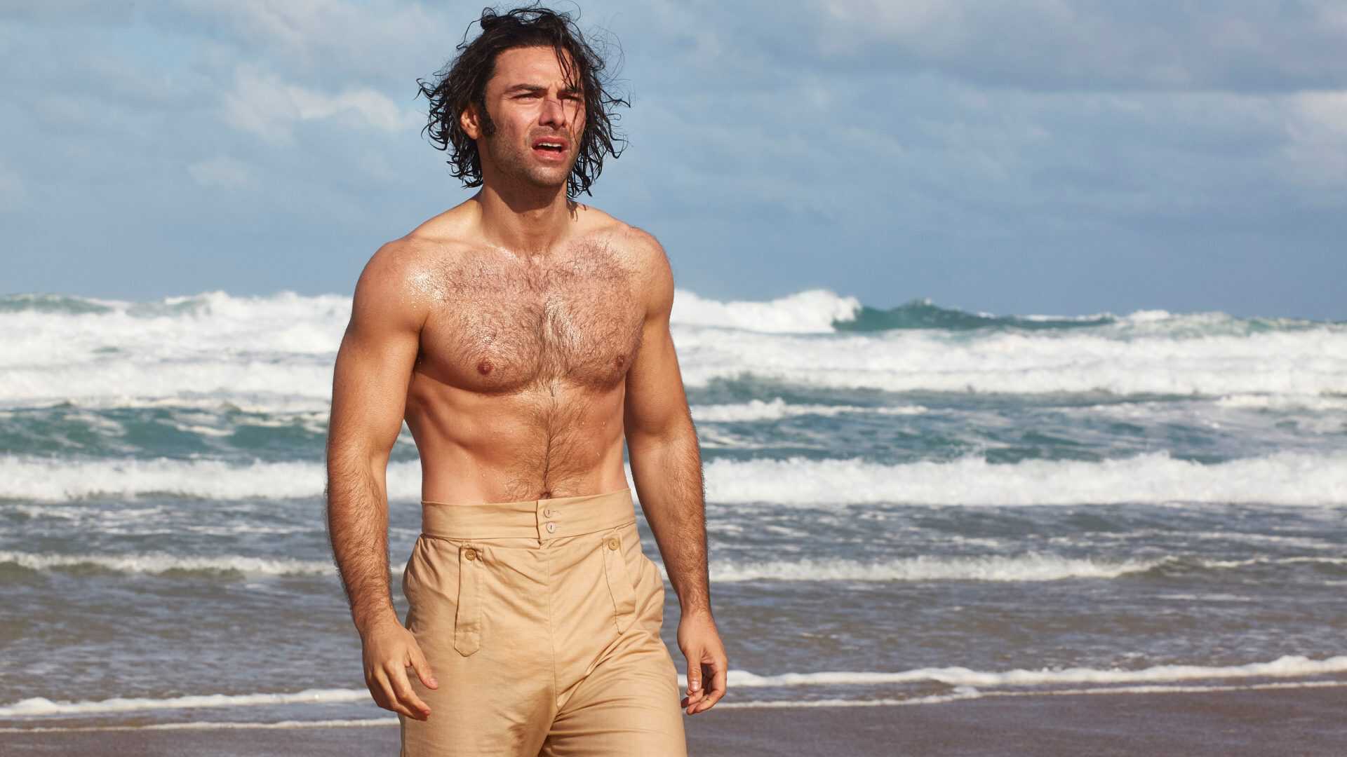 A bear chested man with beige high waist trousers is walking on a beach with waves crashing behind him
