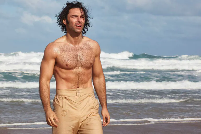 A bear chested man with beige high waist trousers is walking on a beach with waves crashing behind him