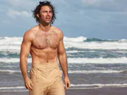 A bear chested man with beige high waist trousers is walking on a beach with waves crashing behind him