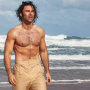 A bear chested man with beige high waist trousers is walking on a beach with waves crashing behind him