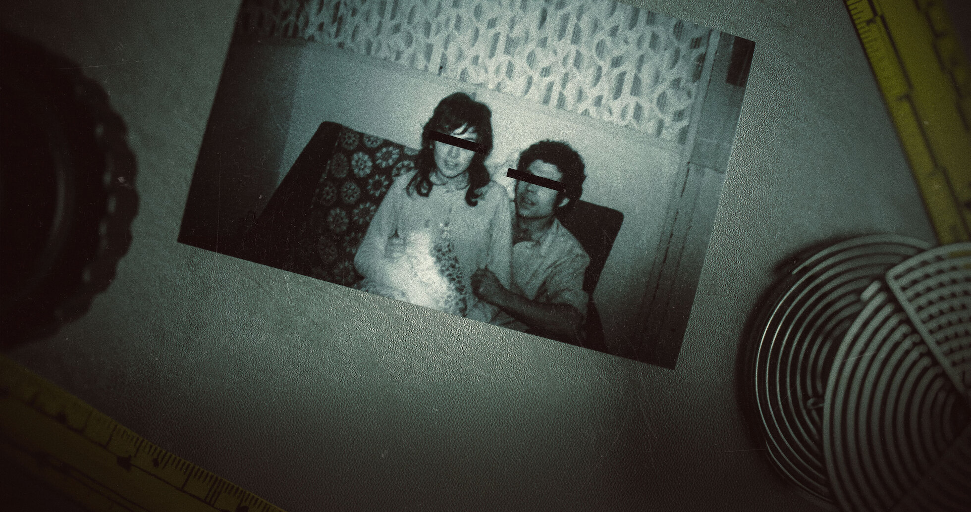 A black and white photo of two people with their eyes crossed out as if redacted sits on a table with