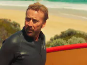 An older man with short hair and a stubble is by a sandy beach wearing a wetsuit and looking concerned as he looks behind him