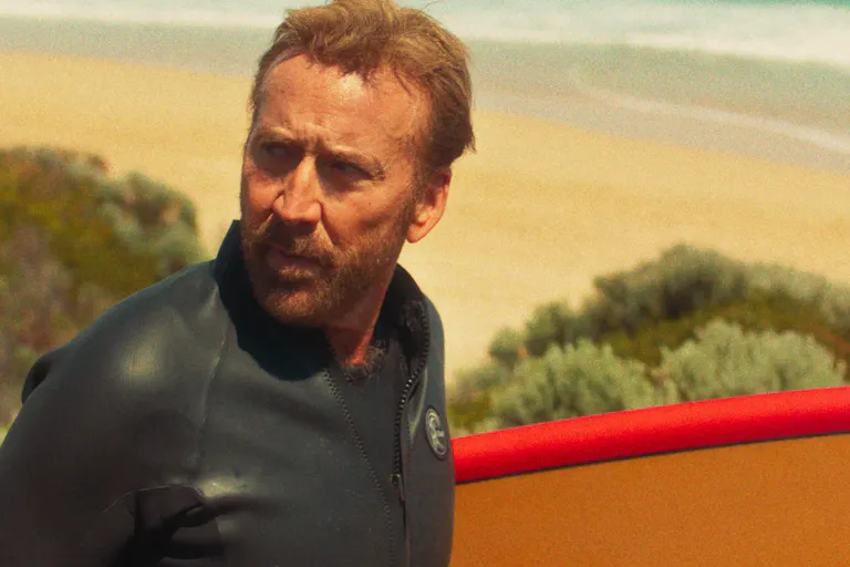 An older man with short hair and a stubble is by a sandy beach wearing a wetsuit and looking concerned as he looks behind him