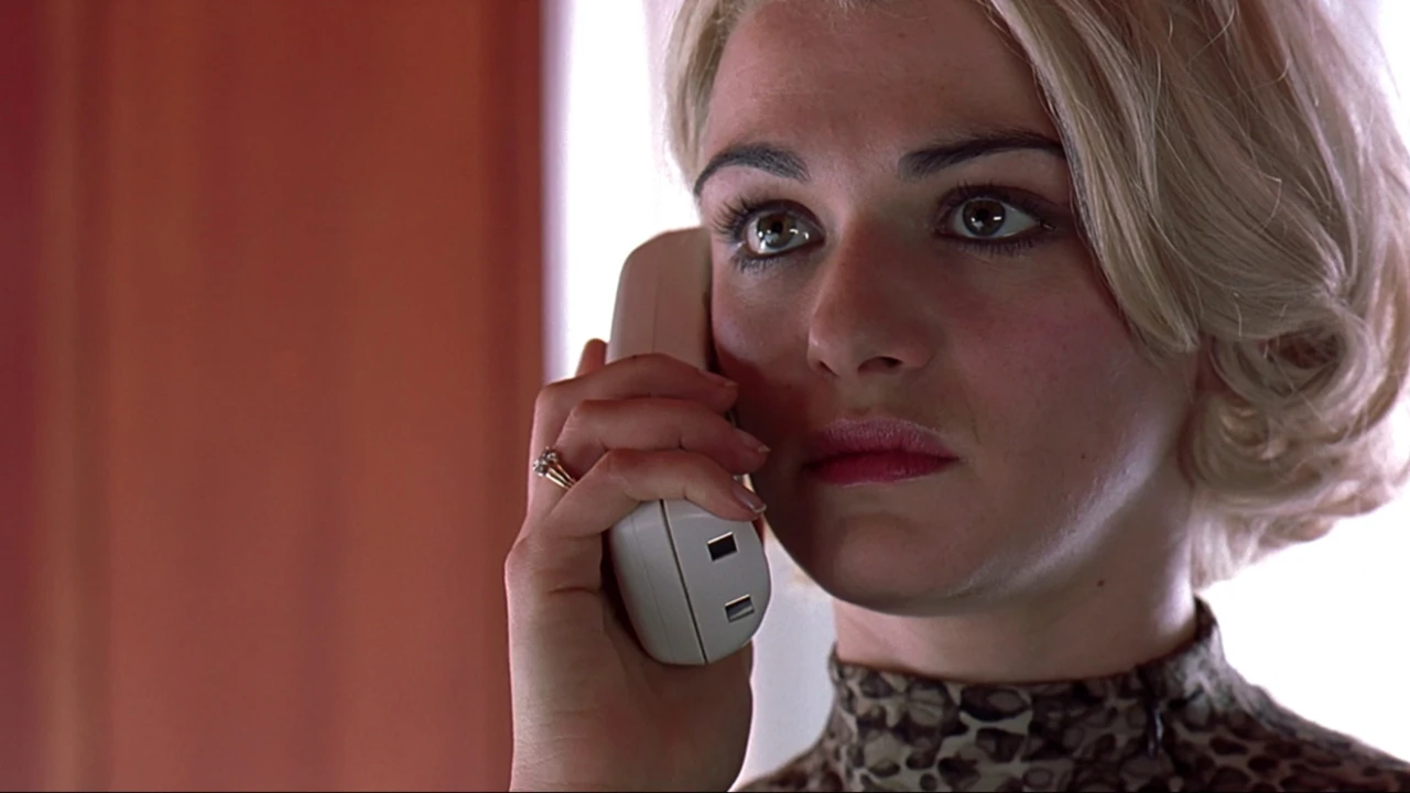 A blond woman with short hair and a determined look is holding a white landline phone to her ear.