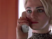 A blond woman with short hair and a determined look is holding a white landline phone to her ear.