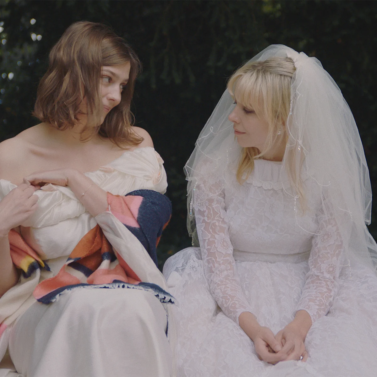 Two women sitting and looking at each other. One is wearing a wedding dress and the other one a formal white dress and a colourful shawl