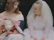 Two women sitting and looking at each other. One is wearing a wedding dress and the other one a formal white dress and a colourful shawl